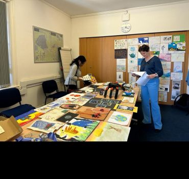 Two women look through submitted artwork on a table. The artworks include 3D sculptures, paintings and collages.