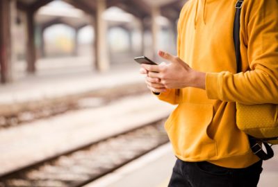 A man dressed in a yellow jumper stands on a train station platform holding a mobile phone in his hands.